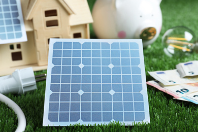 Photo of Solar panels, house model and money on green grass, closeup