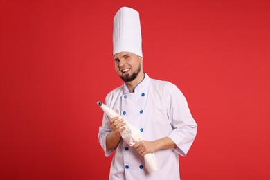 Photo of Happy professional confectioner in uniform holding piping bag on red background