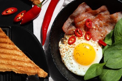 Photo of Tasty fried egg with bacon, chili pepper and spinach on black table, flat lay