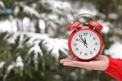 Woman holding red alarm clock near fir tree covered with snow outdoors. Space for text