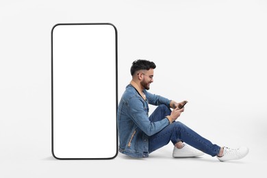 Man with mobile phone sitting near huge device with empty screen on white background. Mockup for design
