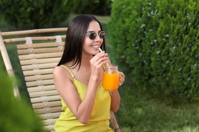Beautiful young woman with refreshing drink relaxing in deckchair at garden