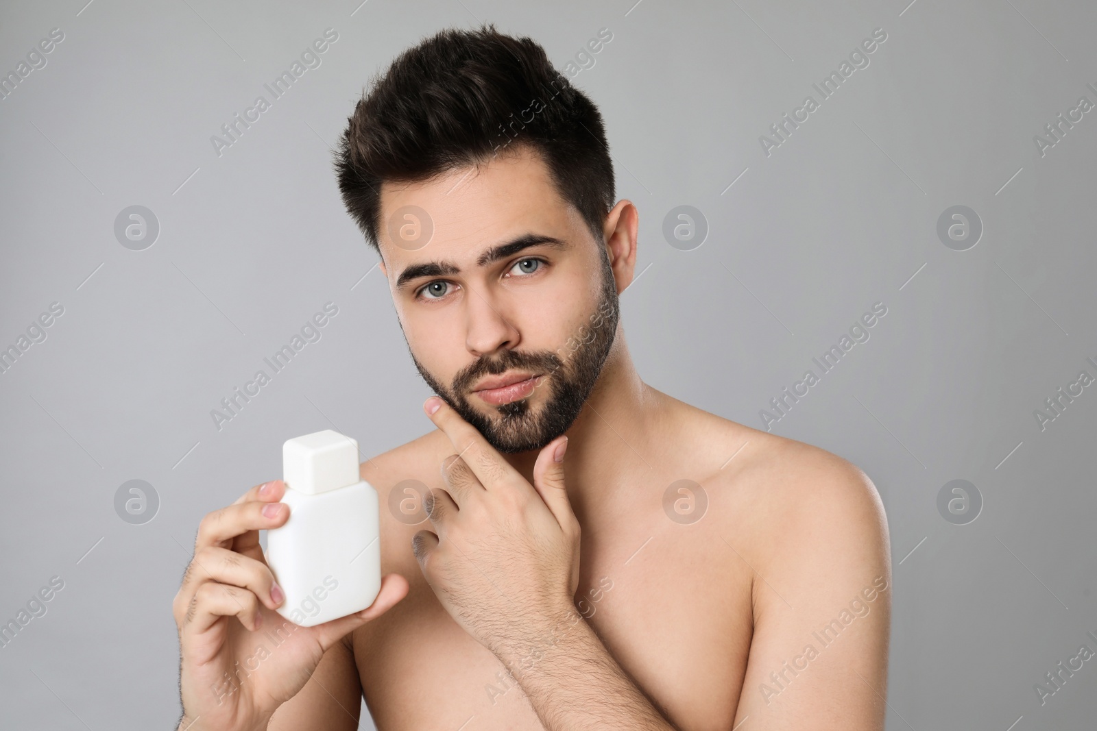 Photo of Handsome young man with beard holding post shave lotion on grey background