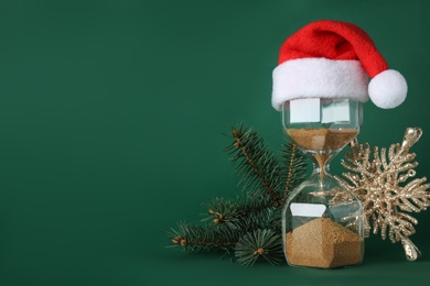 Photo of Hourglass with Santa hat and fir tree branches on green background, space for text. Christmas countdown