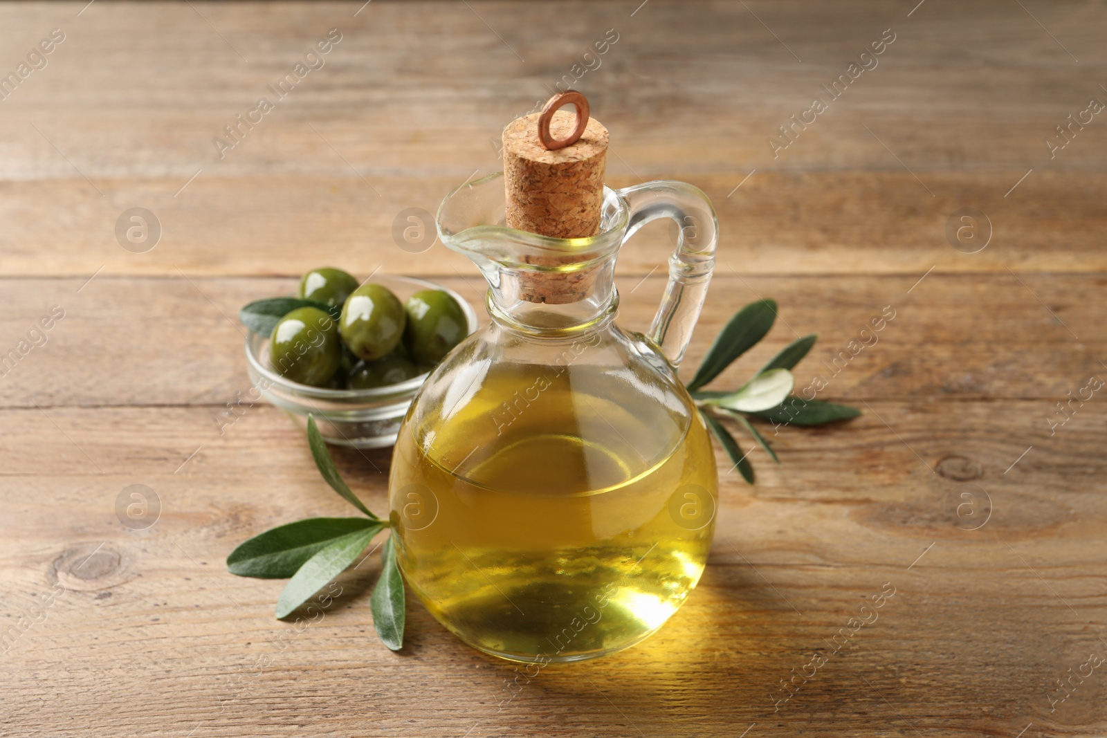 Photo of Glass jug of oil, ripe olives and green leaves on wooden table, closeup