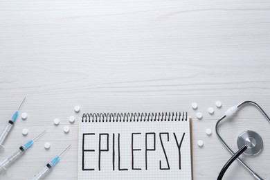 Photo of Notebook with word Epilepsy, stethoscope, pills and syringes on white wooden table, flat lay. Space for text