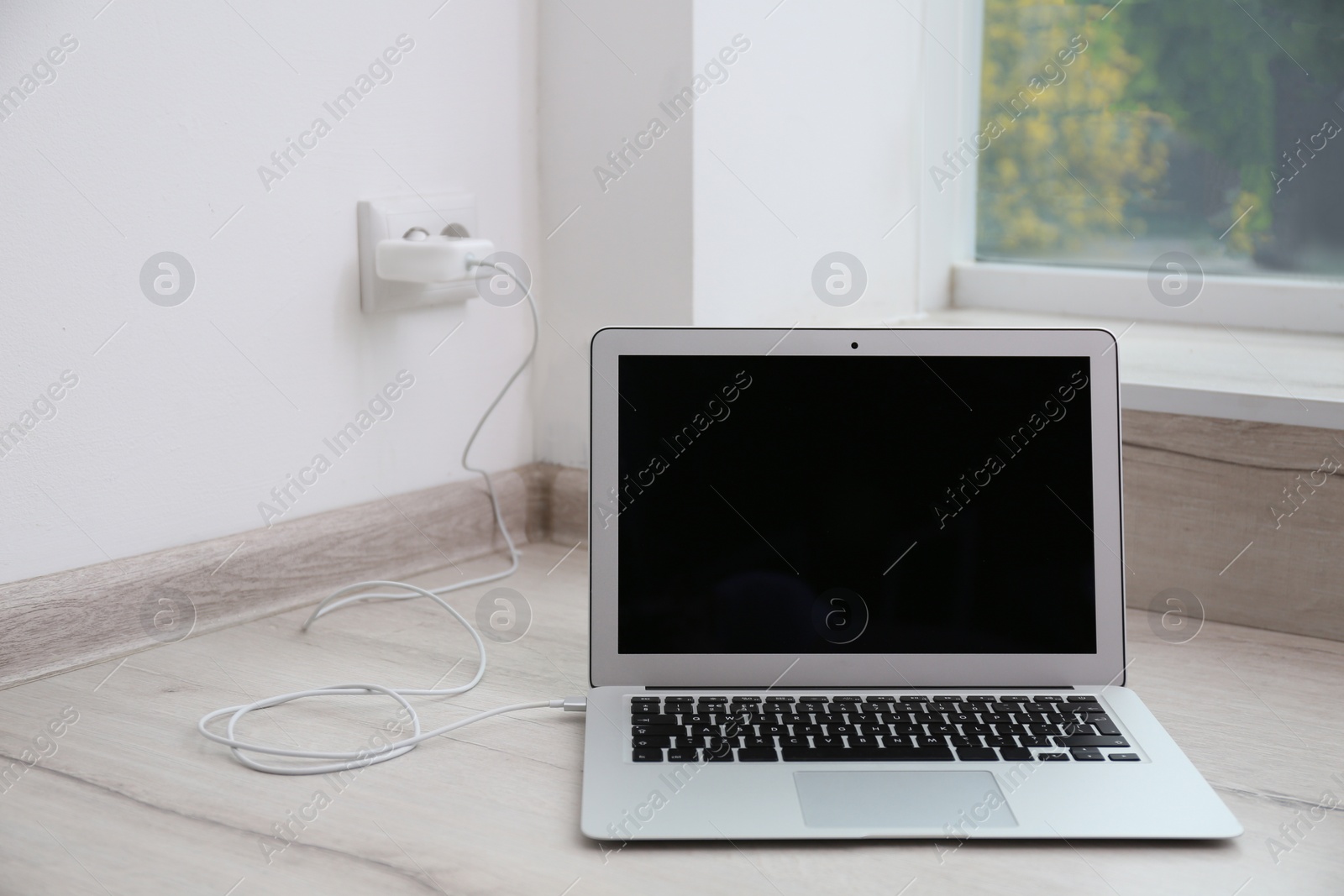 Photo of Modern laptop with charger on floor near white wall