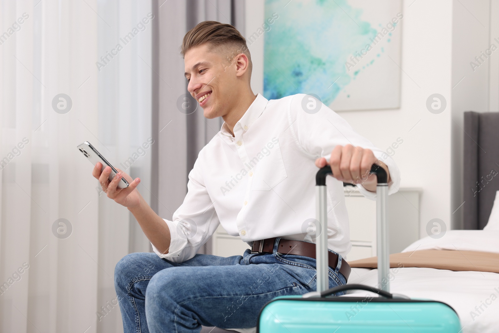 Photo of Smiling guest with suitcase and smartphone on bed in stylish hotel room