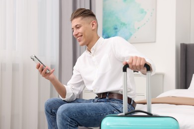 Photo of Smiling guest with suitcase and smartphone on bed in stylish hotel room