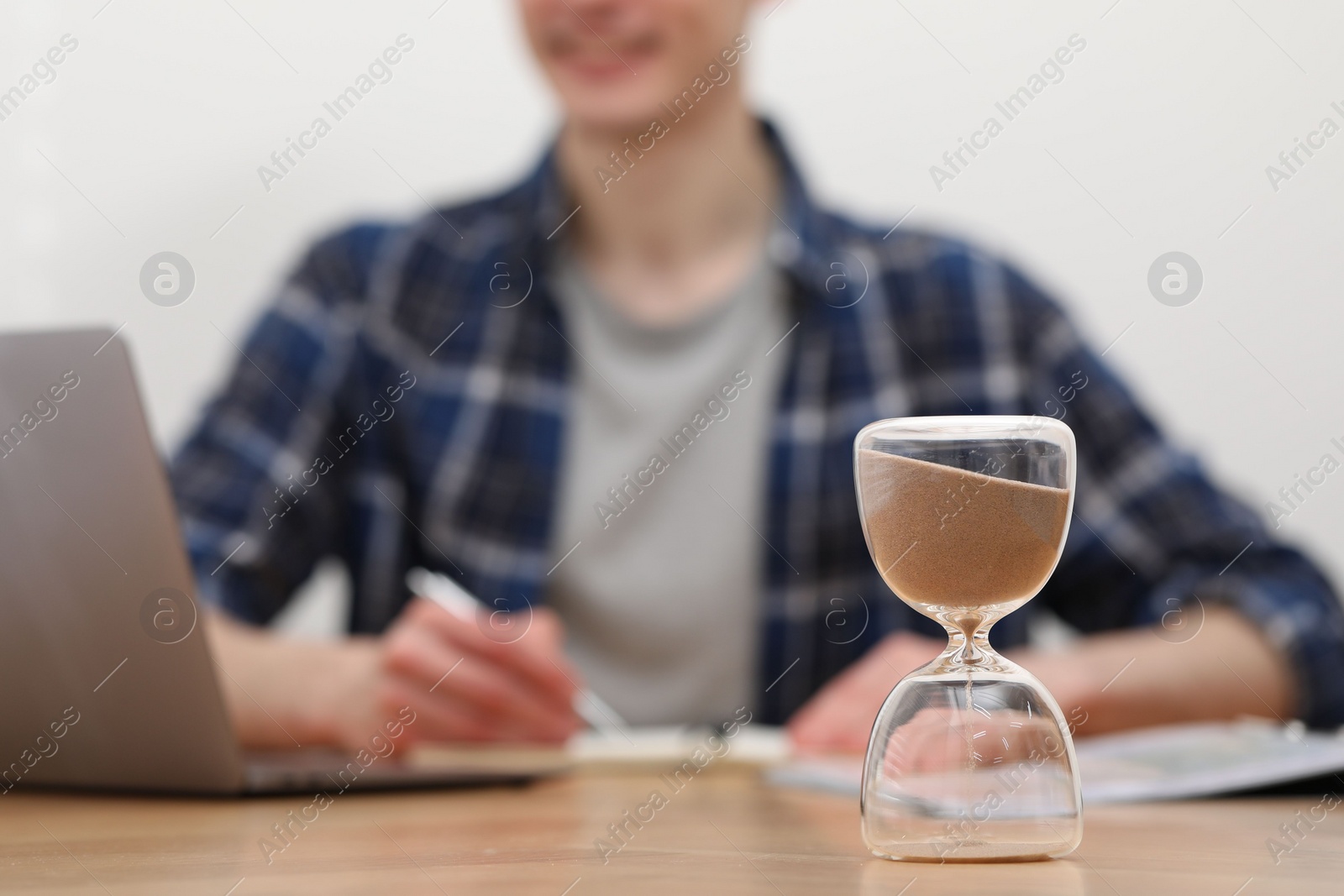 Photo of Hourglass with flowing sand on desk. Man taking notes while using laptop indoors, selective focus