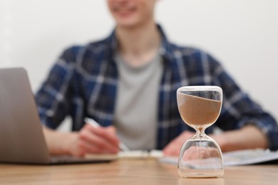 Photo of Hourglass with flowing sand on desk. Man taking notes while using laptop indoors, selective focus