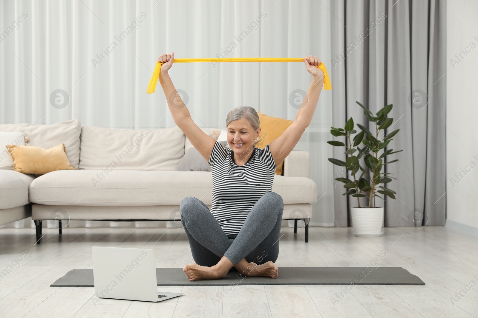 Photo of Senior woman doing exercise with fitness elastic band near laptop on mat at home