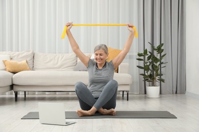 Senior woman doing exercise with fitness elastic band near laptop on mat at home