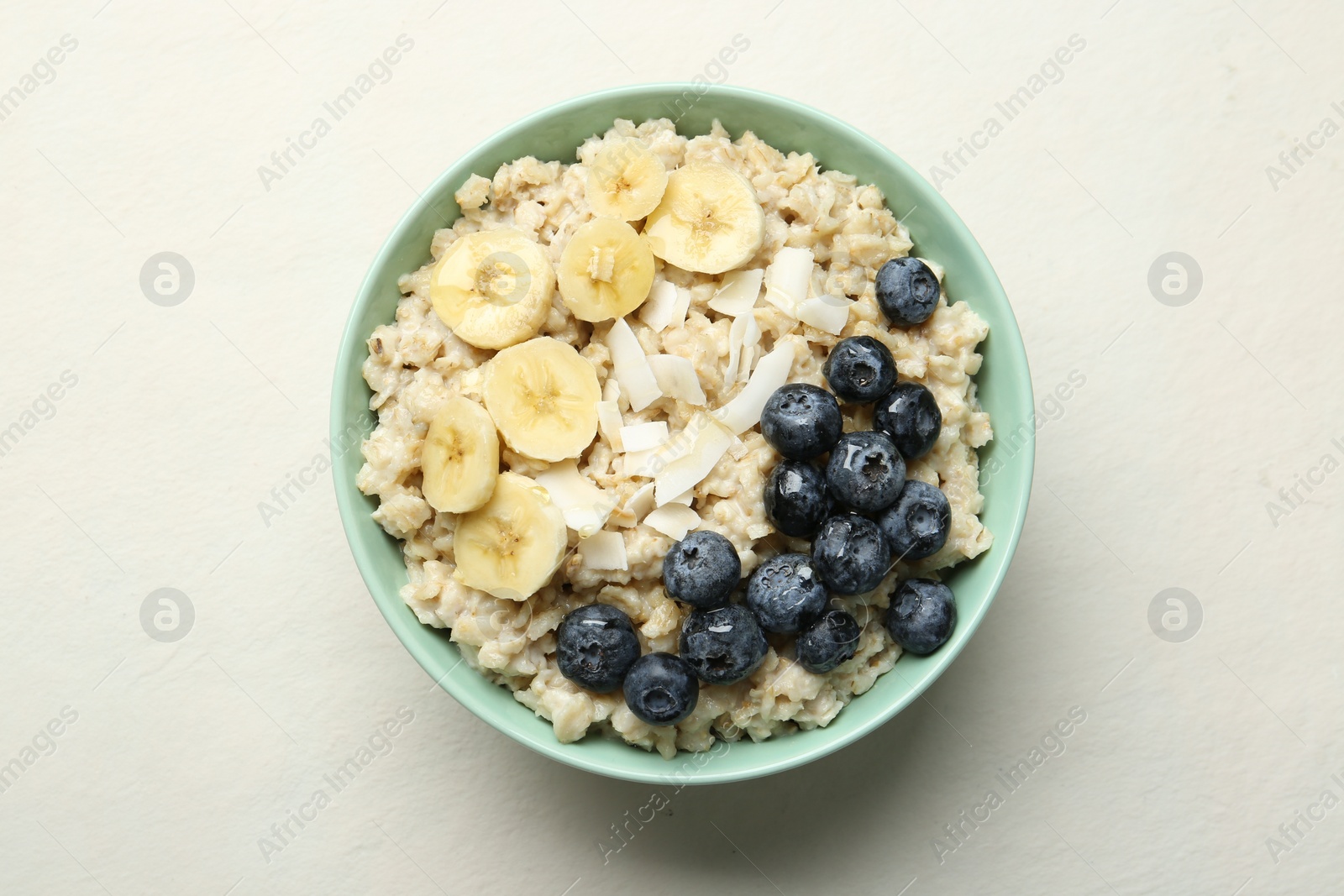 Photo of Tasty oatmeal with banana, blueberries, coconut flakes and honey served in bowl on beige table, top view