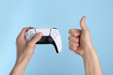 Man using wireless game controller and showing thumbs up on light blue background, closeup