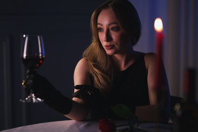 Photo of Elegant young woman with glass of wine at table indoors in evening