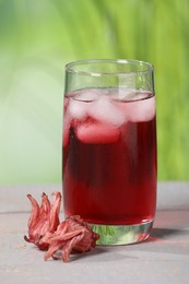 Refreshing hibiscus tea with ice cubes in glass and roselle flowers on white wooden table against blurred green background