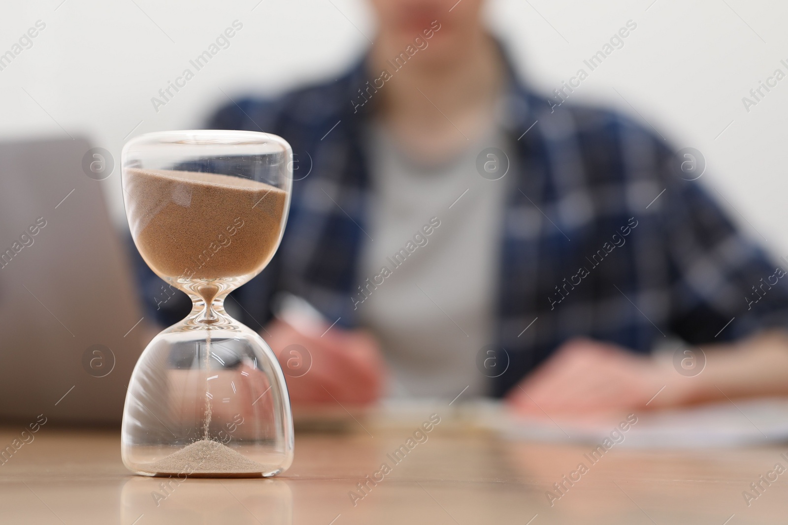 Photo of Hourglass with flowing sand on desk. Man taking notes indoors, selective focus