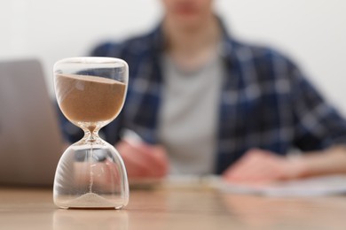 Photo of Hourglass with flowing sand on desk. Man taking notes indoors, selective focus