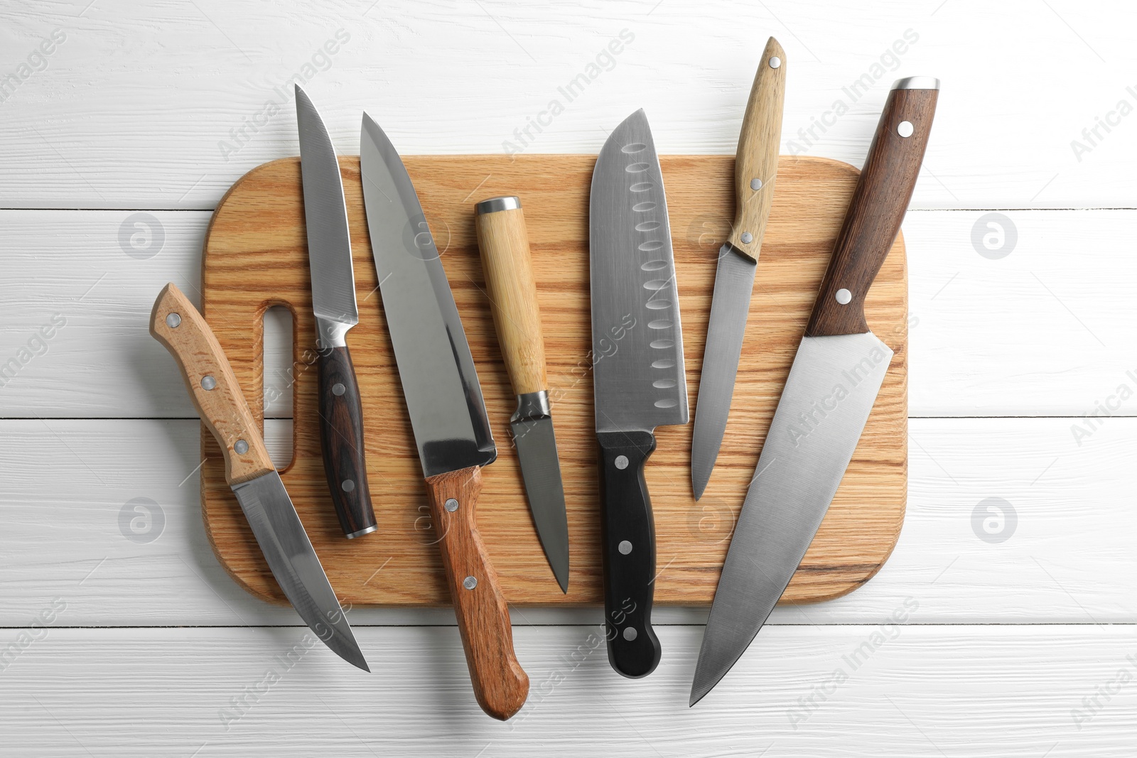 Photo of Many different knives and board on white wooden table, flat lay