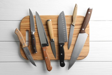 Photo of Many different knives and board on white wooden table, flat lay