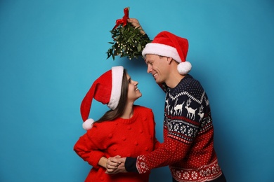 Happy couple standing under mistletoe bunch on light blue background