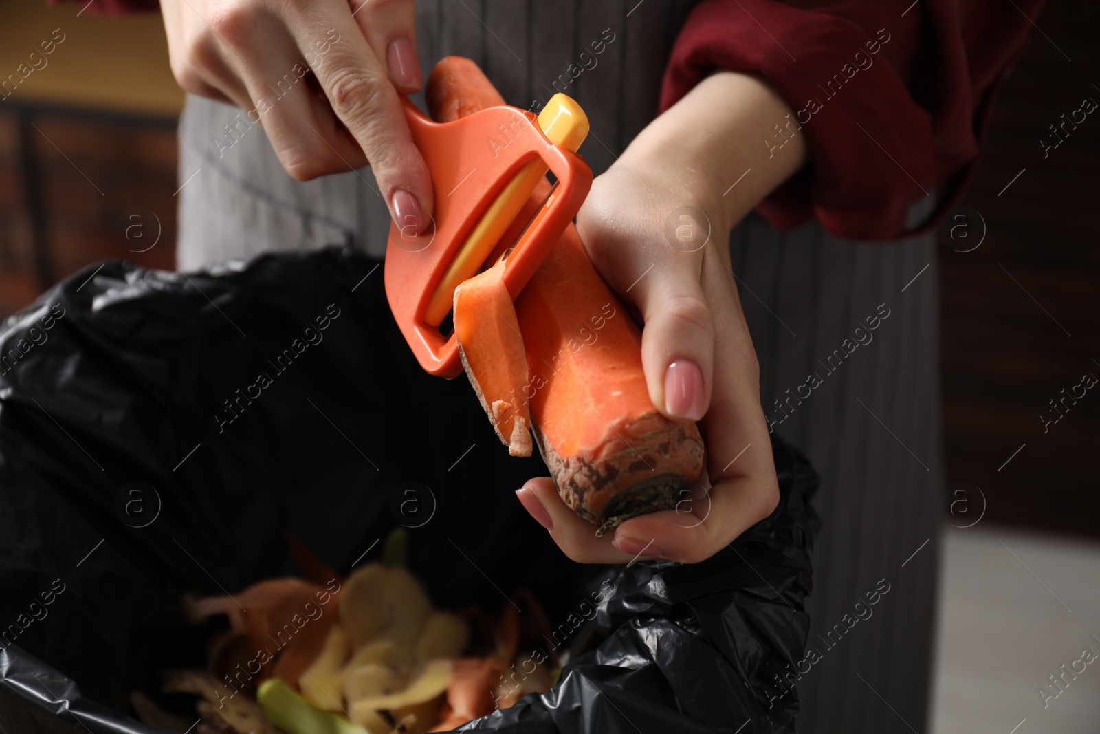 Photo of Woman peeling fresh carrot above garbage bin indoors, closeup