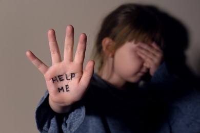 Crying little girl showing palm with phrase HELP ME near beige wall, focus on hand. Domestic violence concept