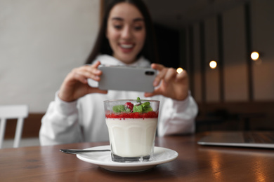 Photo of Young blogger taking picture of dessert at table in cafe, focus on glass
