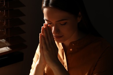 Photo of Religious young woman with clasped hands praying indoors, closeup