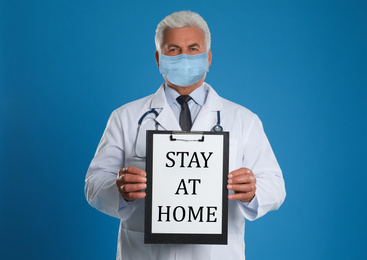 Image of Doctor in medical mask holding clipboard with text STAY AT HOME on blue background