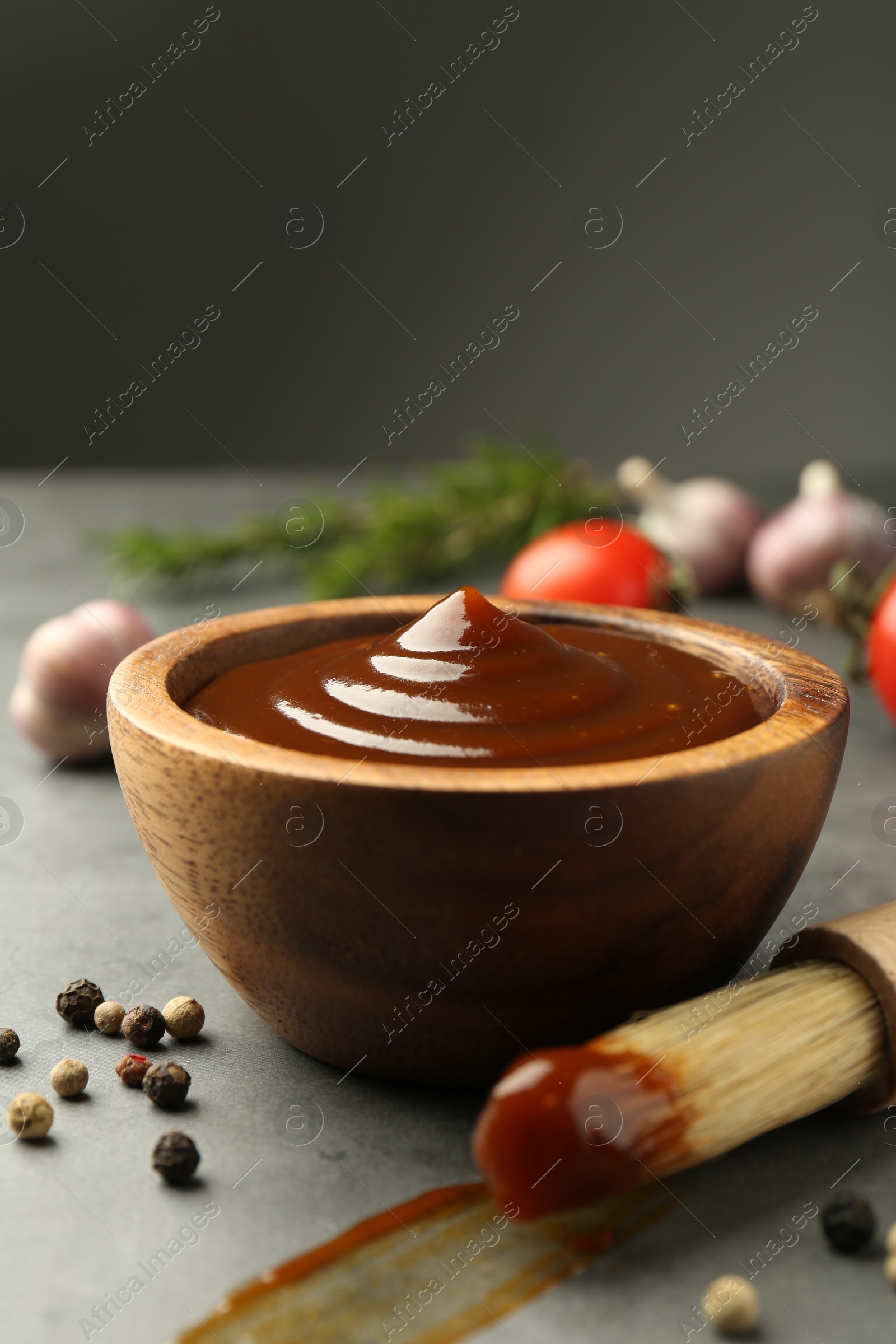 Photo of Tasty barbeque sauce in bowl, brush and peppercorns on grey table, closeup