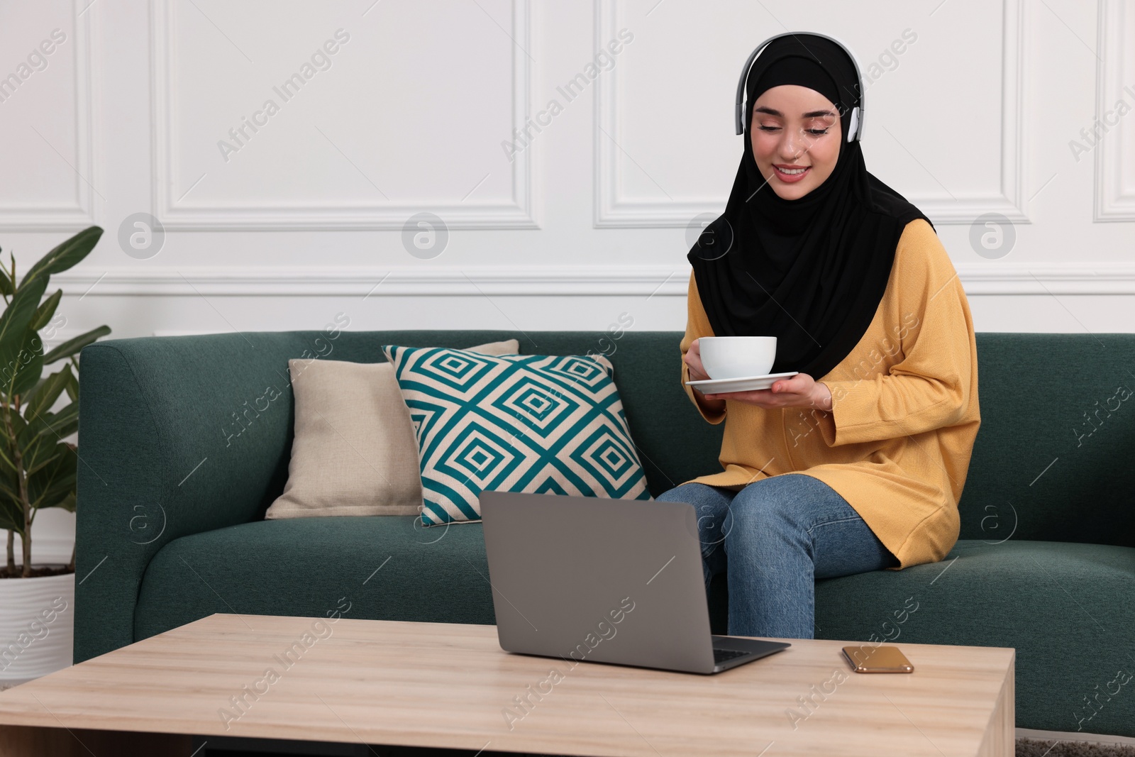 Photo of Muslim woman in hijab with cup of coffee using laptop on sofa indoors. Space for text