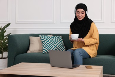 Muslim woman in hijab with cup of coffee using laptop on sofa indoors. Space for text