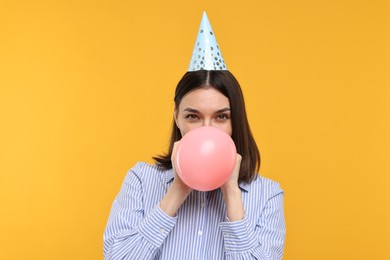 Young woman in party hat blowing balloon on yellow background