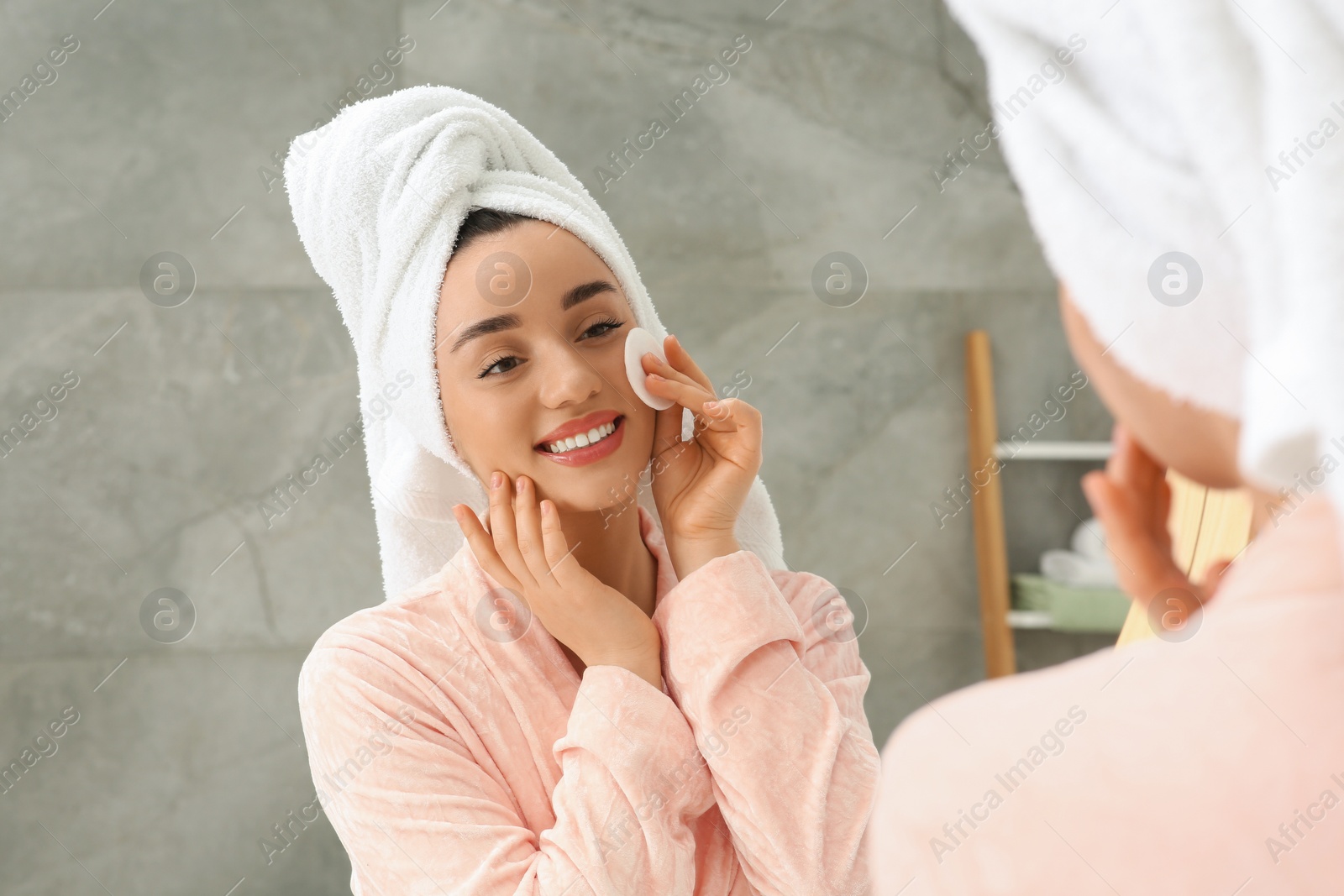 Photo of Beautiful woman in terry towel removing makeup with cotton pad near mirror indoors