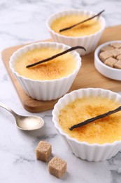 Photo of Delicious creme brulee in bowls, vanilla pods, sugar cubes and spoon on white marble table