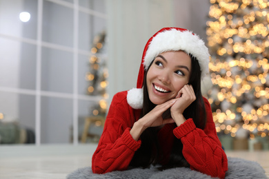 Happy young woman wearing Santa hat in room with Christmas tree