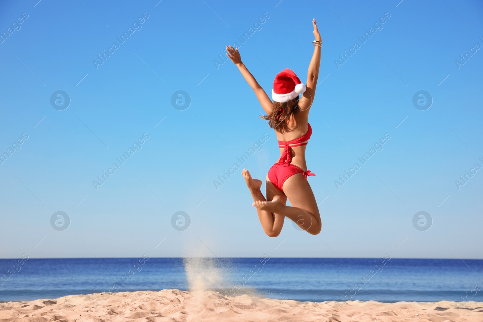 Photo of Young woman in Santa hat and bikini jumping on beach, space for text. Christmas vacation