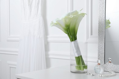 Photo of Beautiful calla lily flowers tied with ribbon in glass vase, bottle of perfume and jewelry on white table. Space for text