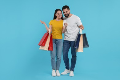 Happy couple with shopping bags and smartphone on light blue background