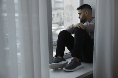 Photo of Sad man sitting on sill and looking at window indoors