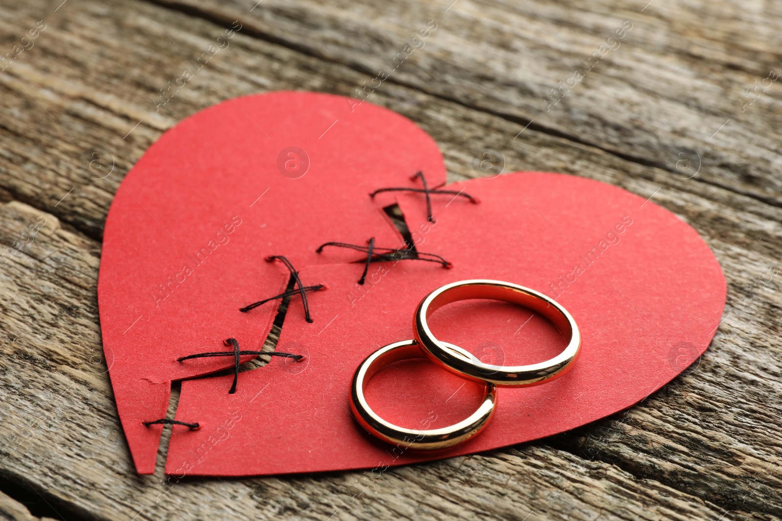 Photo of Broken heart. Torn red paper heart sewed with thread and wedding rings on wooden table, closeup