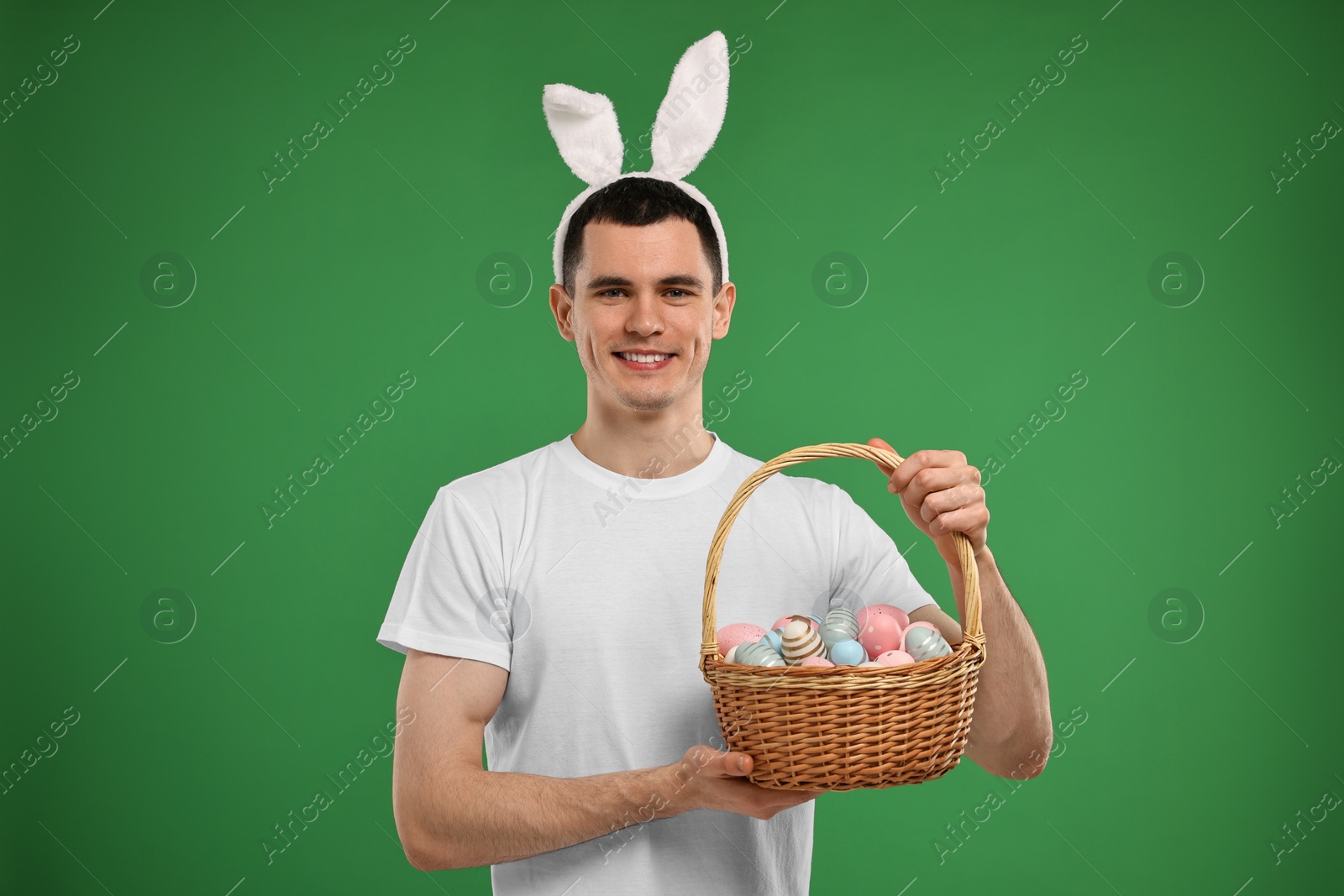 Photo of Easter celebration. Handsome young man with bunny ears holding basket of painted eggs on green background