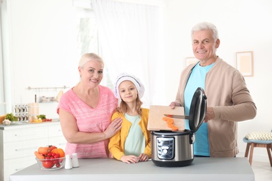 Photo of Mature couple and their granddaughter preparing food with modern multi cooker in kitchen