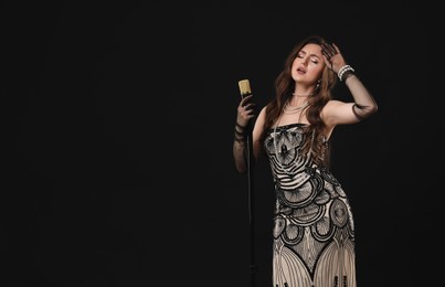 Beautiful young woman in stylish dress with microphone singing on black background, space for text