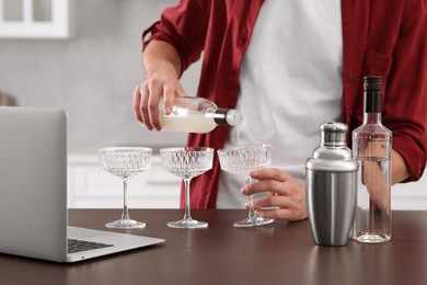 Photo of Man learning to make cocktail with online video on laptop at wooden table in kitchen, closeup. Time for hobby