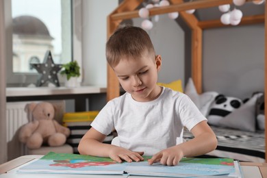 Photo of Cute little boy reading book at table in room