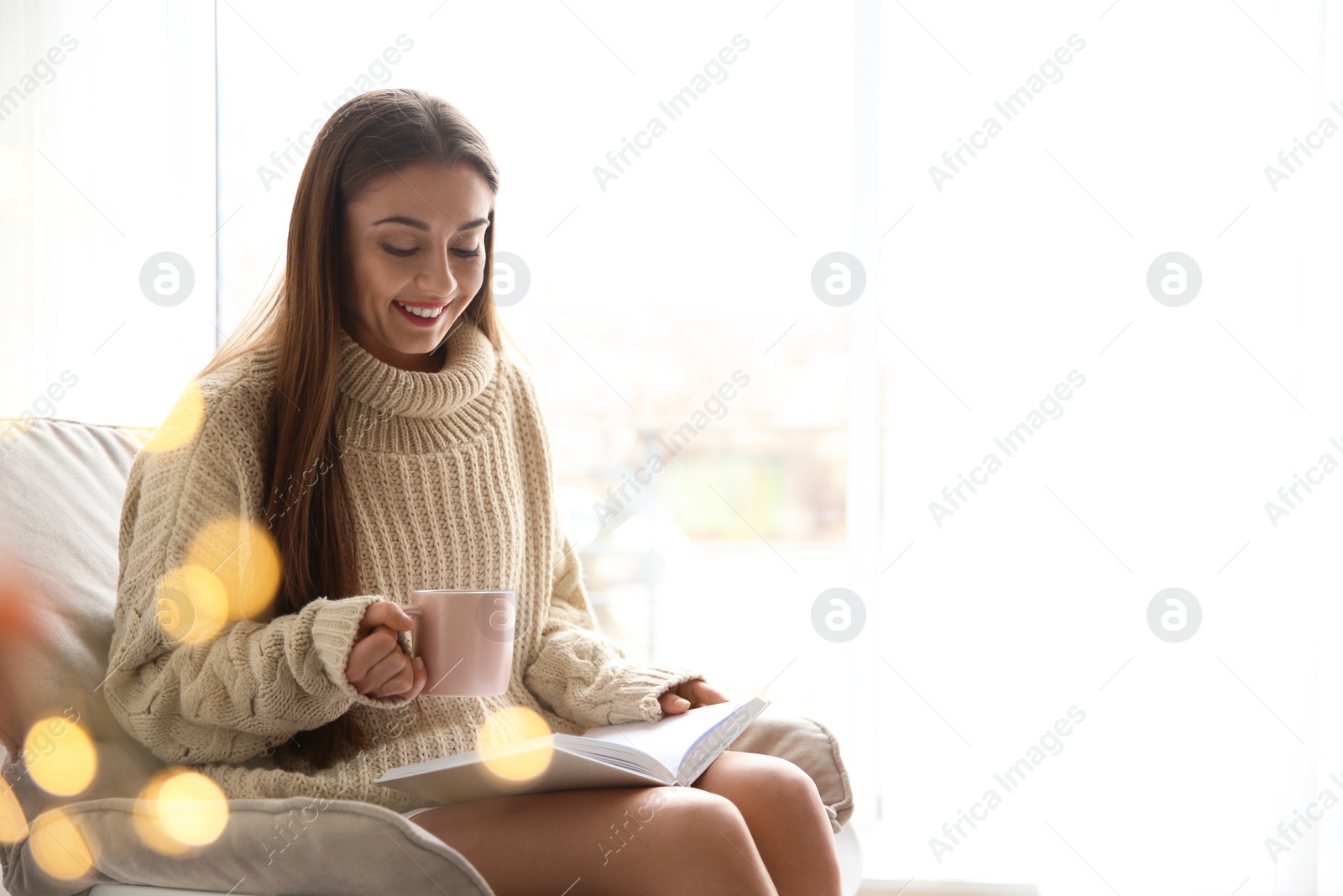 Photo of Young woman with cup of coffee reading book near window at home, space for text