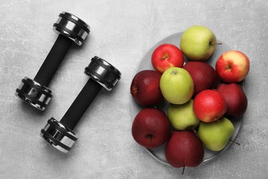 Photo of Healthy diet. Plate with apples and dumbbells on light grey textured table, flat lay